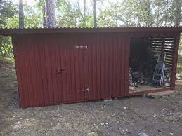 Röd träskjul i skogen, delvis öppen, förvaring av ved, naturskönt, enkelt tak, två dörrar, en stängd, en öppen.