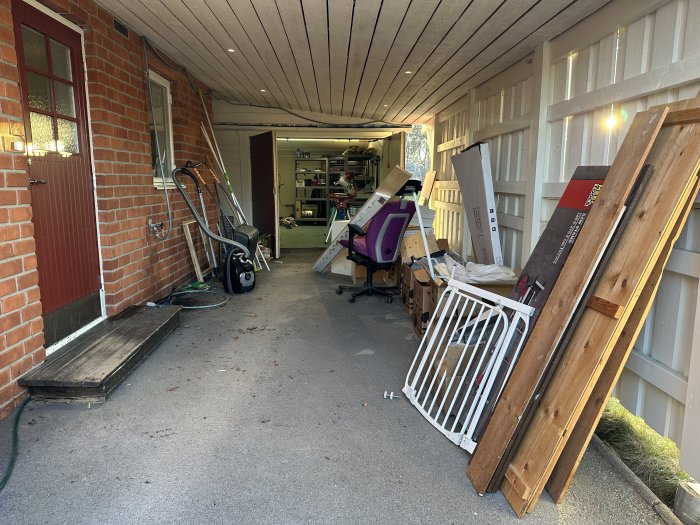 Garage med öppen port och diverse föremål som verktyg och byggmaterial. Röd tegelbyggnad, trästaket och soligt väder.