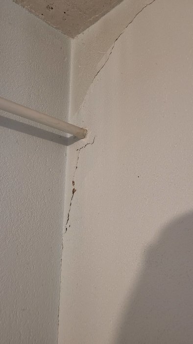 Vägg med spricka vid taket, grov textur, rör längs väggen, skuggor, skador eller slitage synligt.