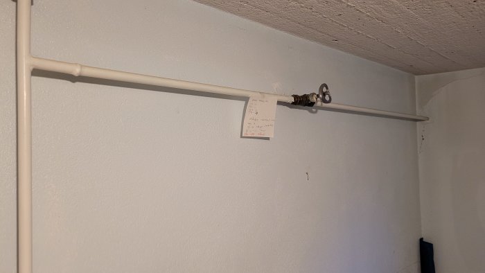 Vit rörledning längs vägg, lapp med anteckningar, vattenavstängningsventil, intetsägande interiör, sprickor på vägg.