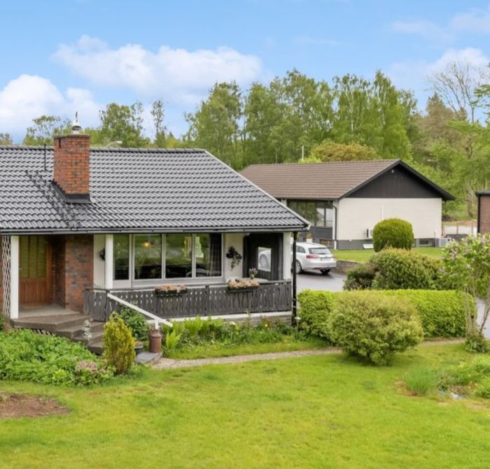 Ett enplanshus med veranda, tegelskorsten, grönskande trädgård, grannhus i bakgrunden, lugnt bostadsområde.