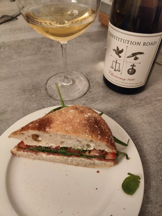 Halverad sandwich med tomater och grönsaker, ett glas vitt vin, vinflaska med etiketten "Constitution Road Chardonnay 2020".