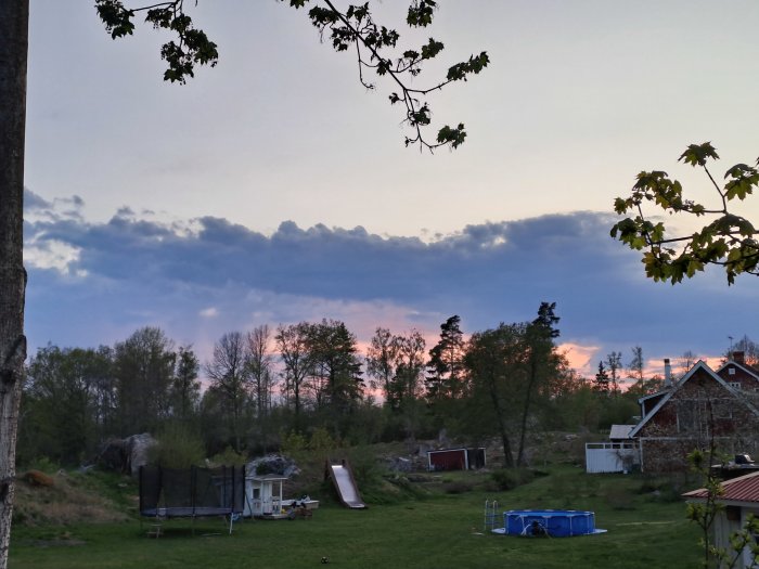 Skymning över en grönskande trädgård med studsmatta, lekstuga, rutschkana och en oanvänd pool. Dramatiska moln i bakgrunden.