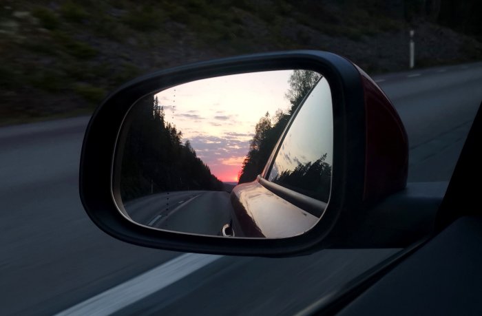 Bilens sidospegel reflekterar en solnedgång och en tom väg omgivna av skog.