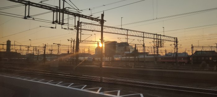 Solnedgång vid en järnvägsstation med tåg, kontaktledningar, och stadssiluett i bakgrunden.