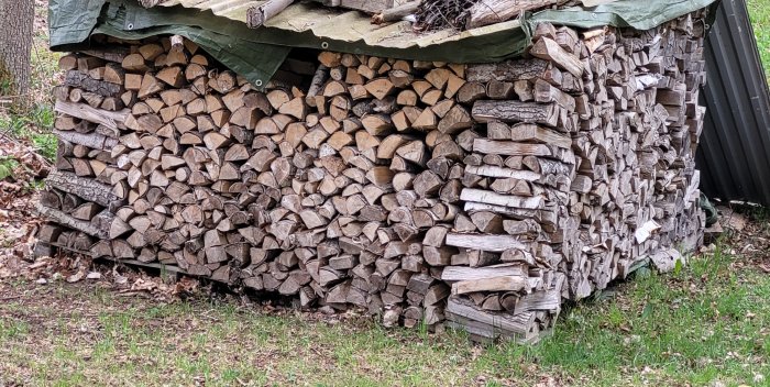 Vedstapel utomhus täckt med presenning, arrangerad mot ett skjul.