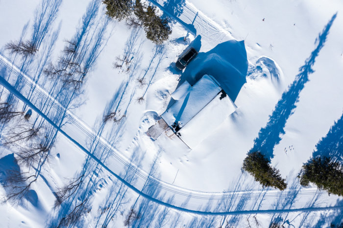 Flygbild över snötäckt landskap med hus, träd, skuggor och en bilvy.