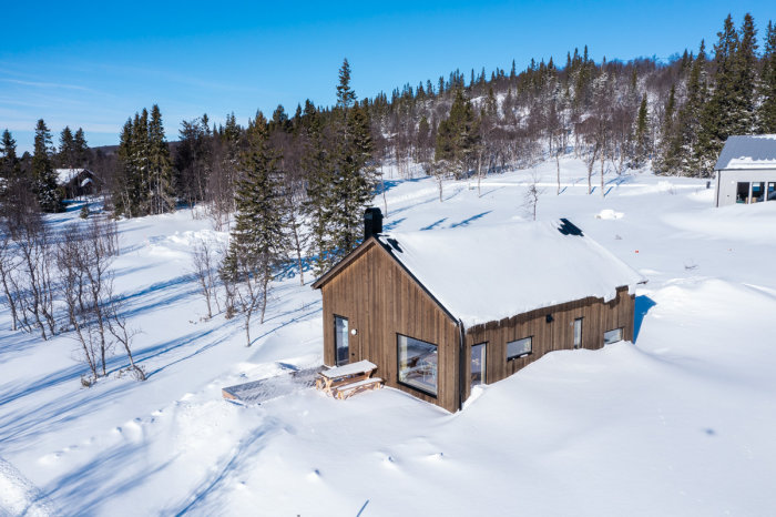 En trästuga i ett snöigt landskap, omgiven av träd under klarblå himmel.