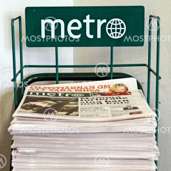 En hög med gratis dagstidningar "Metro", staplade i en grön ställning, vägg bakom.