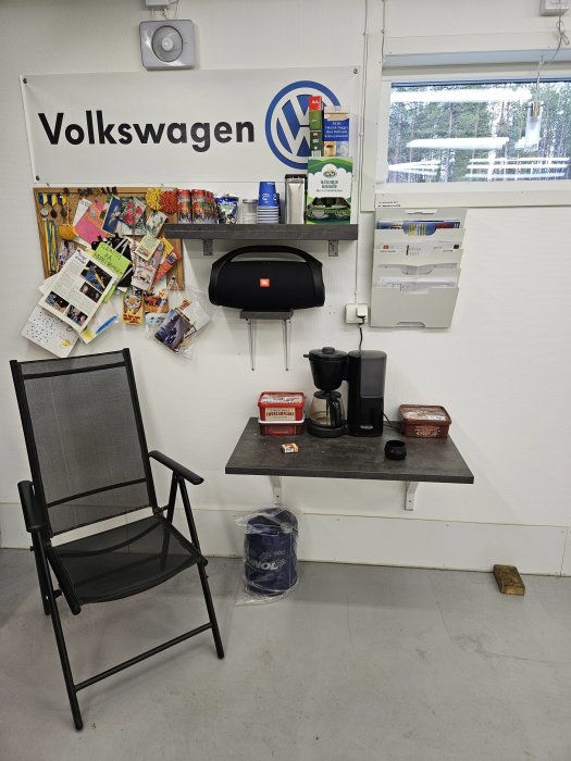 Pausrum med Volkswagen-skylt, kaffemaskin, snacks, stol och sopor. Inomhus, enkelt och funktionellt.