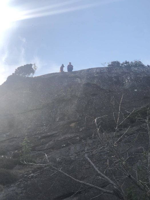 Två personer och en hund på en klippavsats, omgivna av natur, under en ljus himmel.