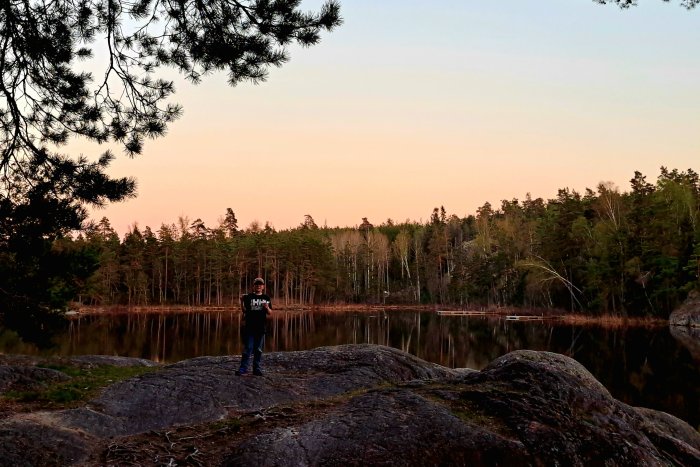 Person står på klippa vid skogssjö i skymning, tallgrenar ramar in bilden överst.