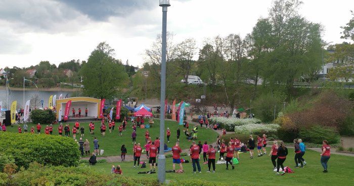 Utommhusaktivitet, träning, grupp människor i röda tröjor, tält, banderoller, grönområden, molnig himmel.