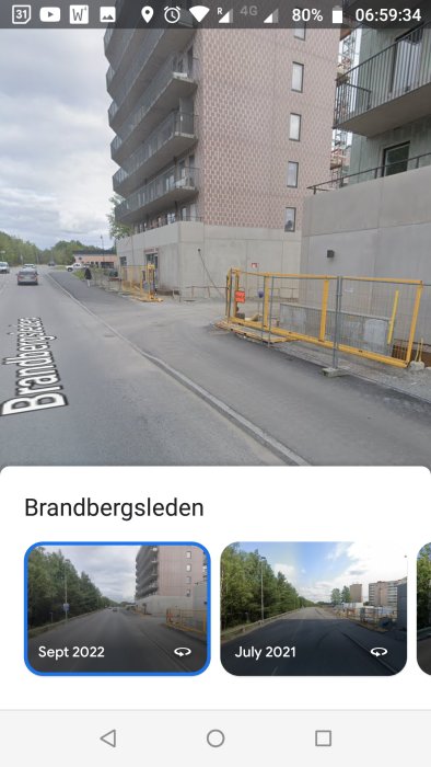 Stadsmiljö med höghus, vägarbeten, stängsel, fordon och asfalterad gata. Skärmbilden tyder på tidsförändring.