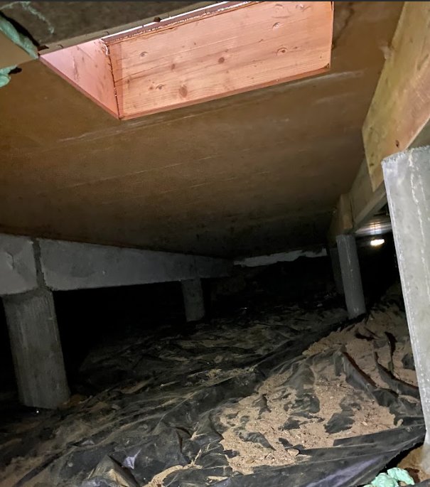 Mörkt kryputrymme under hus med trästöd, plast och isolering. En öppning syns ovanför.