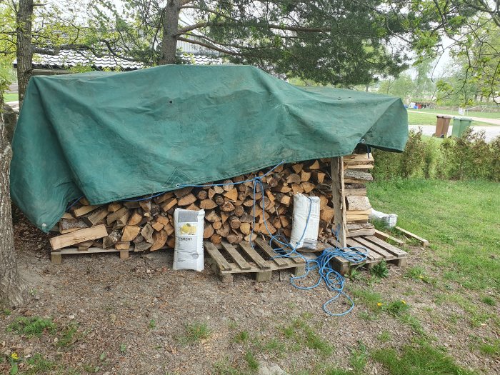 Staplade vedträn skyddade av grön presenning, säckar, rep, utomhusmiljö.