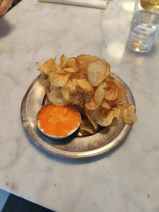 Potatischips på tallrik med dippsås på marmorbord. Snacks och vattenglas i bakgrunden.