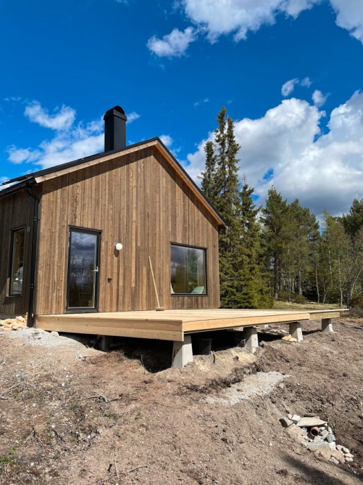 En stuga med träfasad, altan, skorsten under en blå himmel och moln, omgiven av skog.