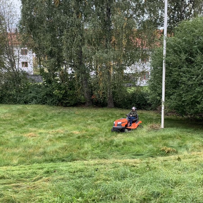 Person kör röd gräsklippare på ojämnt grönt gräs, träd och vit byggnad i bakgrunden.