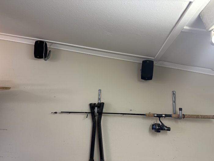 Fiskespö upphängt på vägg med krokar, två högtalare monterade på taket, inomhusbelysning, neutral väggfärg.