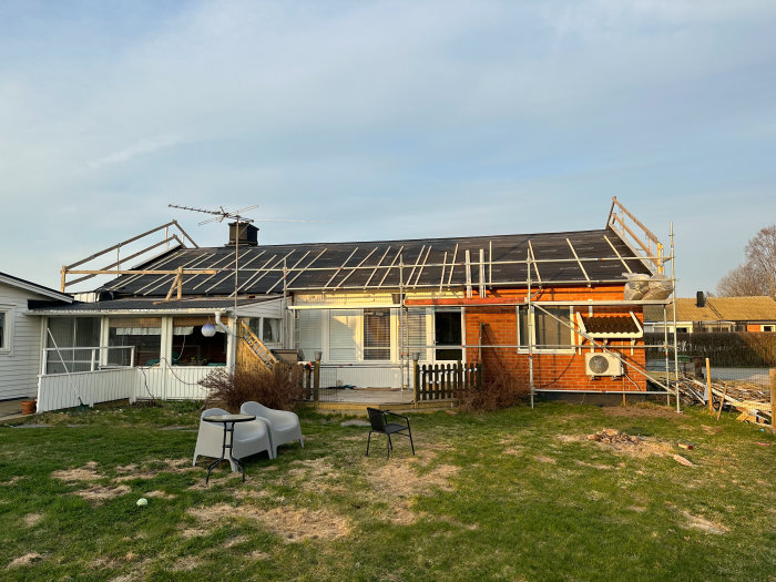 Hus med pågående takarbete, byggställningar, gräsmatta, trädgårdsmöbler, klart väder.