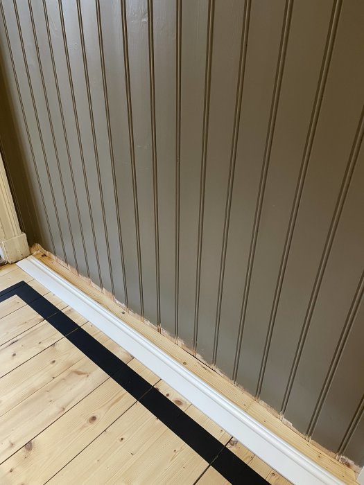 Trägolv, vit golvlister, rutigt mönster, brun vägg med vertikala paneler. Inredningsdetaljer i ett rum.