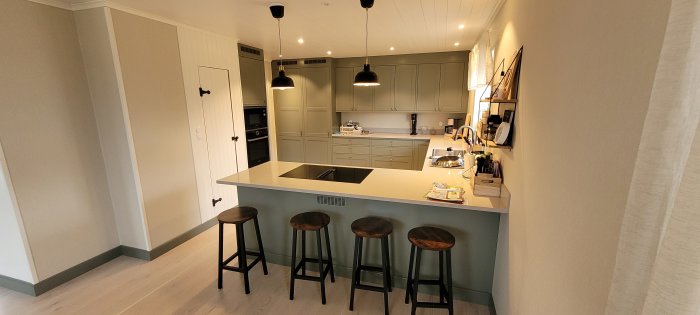 Modernt kök med köksö, pallar, inbyggda apparater, grå skåp, svart armatur och hängande lampor.