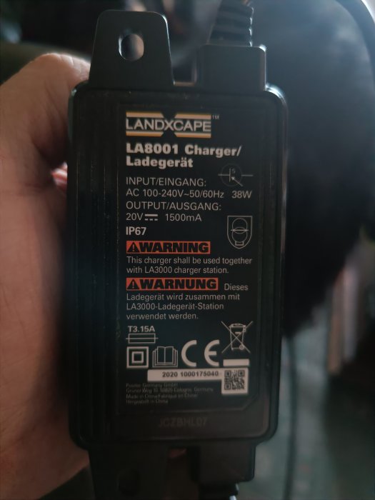 Hand håller svart LANDXCAPE batteriladdare. IP67, 38W, 20V-1500mA utdata. Varningstexter på engelska och tyska.