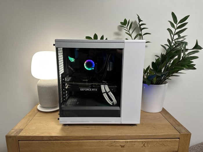 En dator med öppet chassi, RGB-belysning, på ett träbord, intill växter och en lampa.