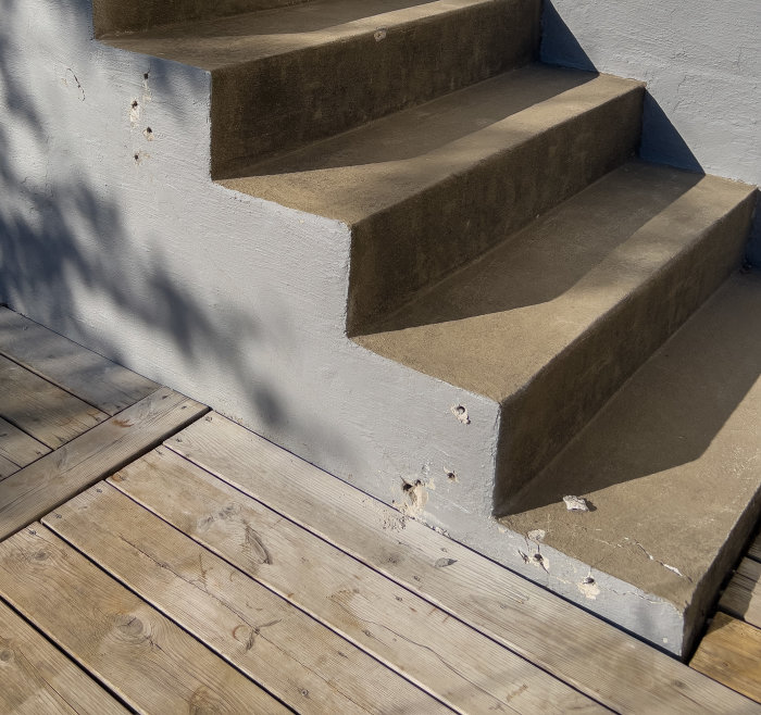 Trägolv möter betongtrappa med skuggor. Solbelyst, utomhus, sliten, vardaglig.