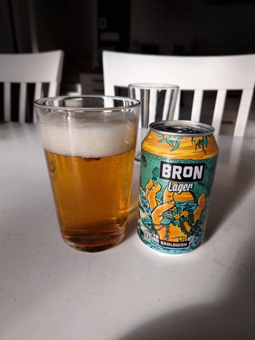 Ett glas öl bredvid en färgglad ölburk märkt "BRON Lager" med texten "Ekologisk" på ett vitt bord.