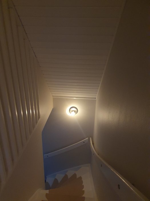 Trappa uppåt med vita väggar, taklampe, träräcke, sett från nedre trappsteg.