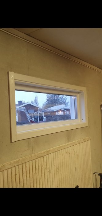 Utsikt genom fönster; grävmaskin och hus utomhus; inomhusmiljö med radiator och vägg.