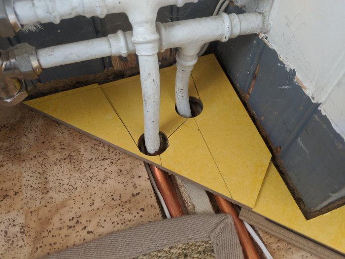 Radiatorrör genom golv, gula isoleringsplattor synliga, oavslutat byggarbete, bruna specklad golvbeläggning.