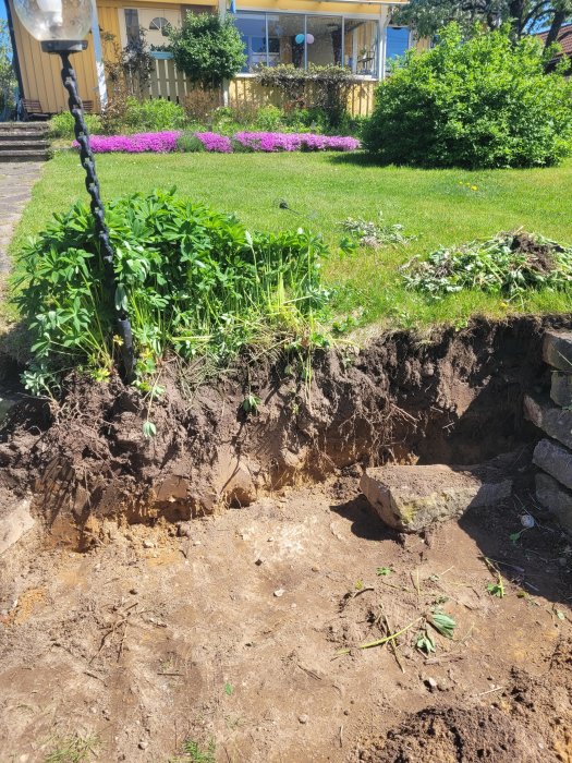 Jordskred nära grönskande trädgård och hus, synlig erosion och trädrotor, solig dag.