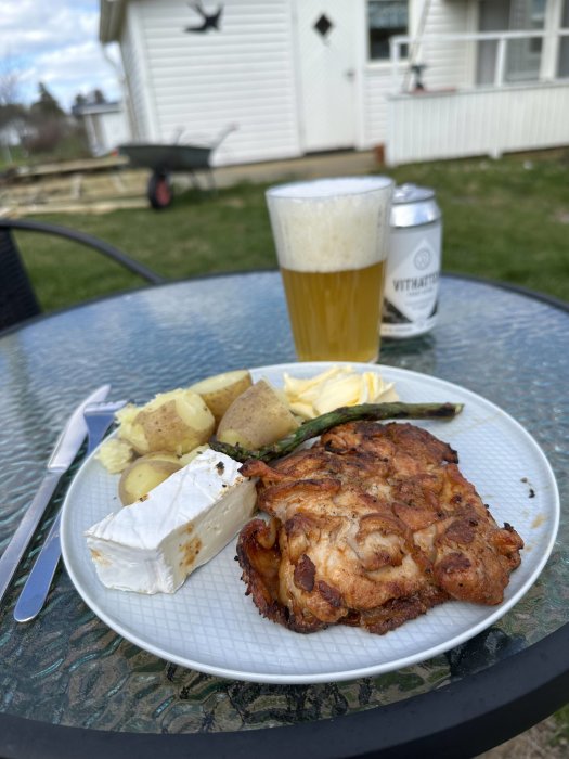 Måltid utomhus med grillad kyckling, potatis, brieost, sparris, öl och gräsklippare i bakgrunden.