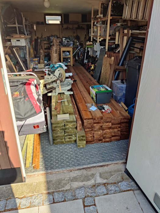 Verkstad full med verktyg, trä, och cyklar. Arbetsbänkar. Rörigt utrymme. Snickeri eller renoveringsprojekt pågår.