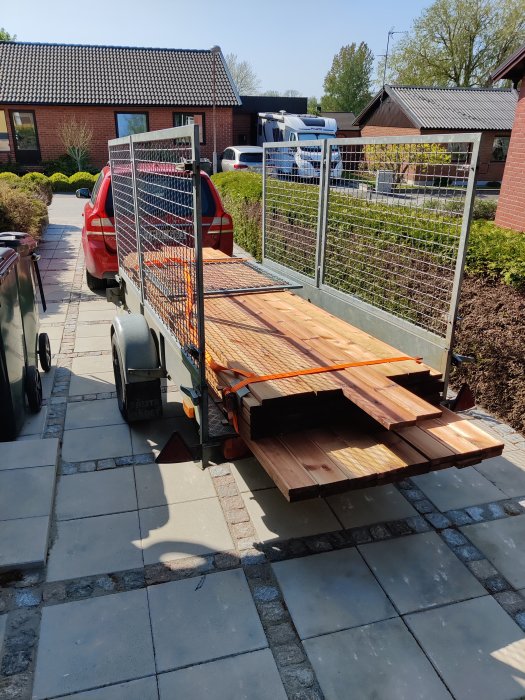 Släpvagn lastad med träbrädor, röd bil, soligt vädret, stenlagd uppfart, bostadsområde.