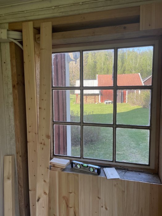 Gammalt fönster i renoveringsfas, utsikt över hage och röda byggnader, vattenpass på fönsterbräda, träväggar.