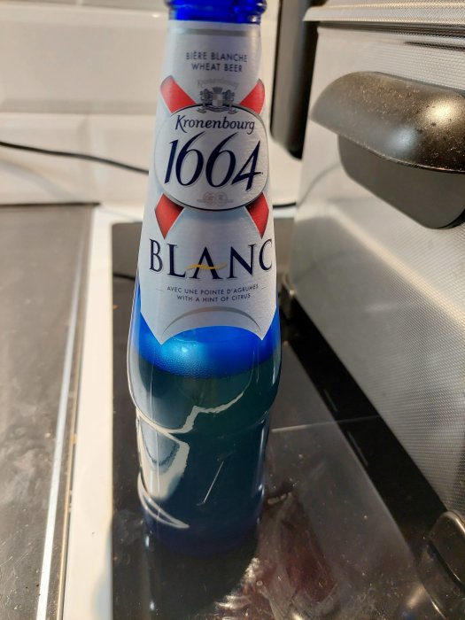 En flaska Kronenbourg 1664 Blanc veteöl med blå kork, på en köksbänk.