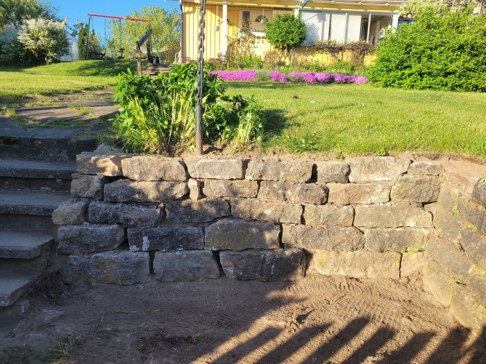 Trädgård med stenmur, trappsteg, grönt gräs, växter och gult hus i bakgrunden under soligt väder.