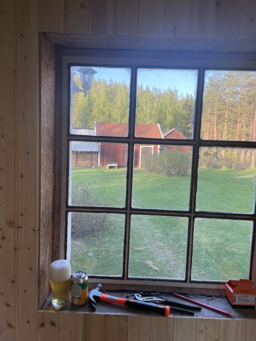 Träfönster med utsikt över trädgård, verktyg och öl på fönsterbräda. Lantlig stämning, avkoppling efter arbete.