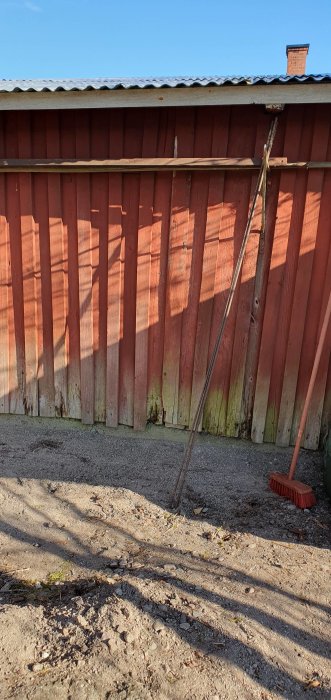 Röd trävägg, skuggor av träden, kvastskaft lutat mot väggen, friluft, grovt grus.