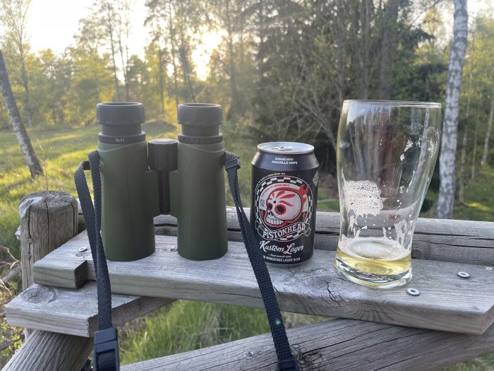 Kikare på träbänk, ölburk, glas med ölrester, naturbakgrund vid solnedgång.