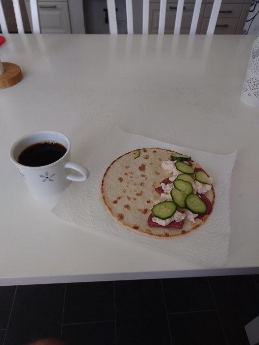 En kopp kaffe och en tunnbrödsrulle med pålägg på ett vitt köksbord.