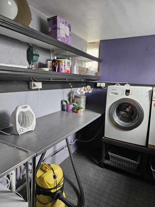Tvättstuga med tvättmaskin, hyllor, städsaker och en värmefläkt. Violett vägg, grå bänkskiva och golv.