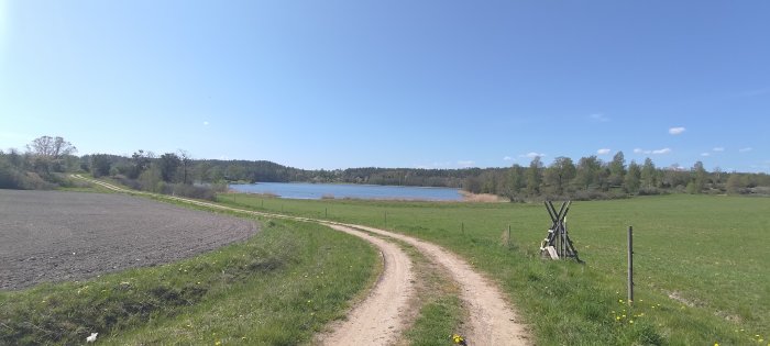 Lantlig stig bredvid plöjd åker, grönt fält, blå himmel, skog, sjö i fjärran. Ljus, fridfull, vårdag.