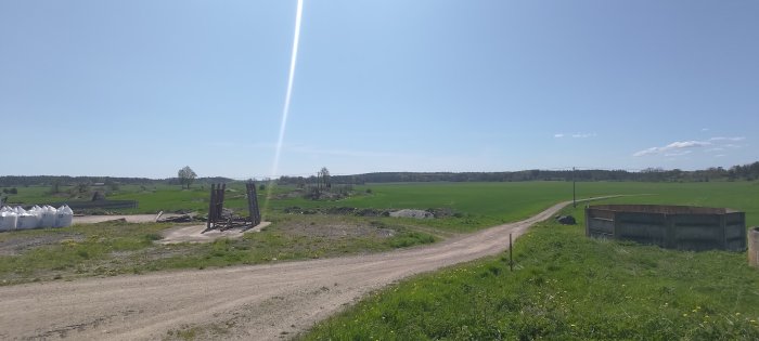 Solig dag, grusväg, gröna fält, lantligt, anläggningsmaterial, träd, blå himmel, glesbebyggelse i bakgrunden.