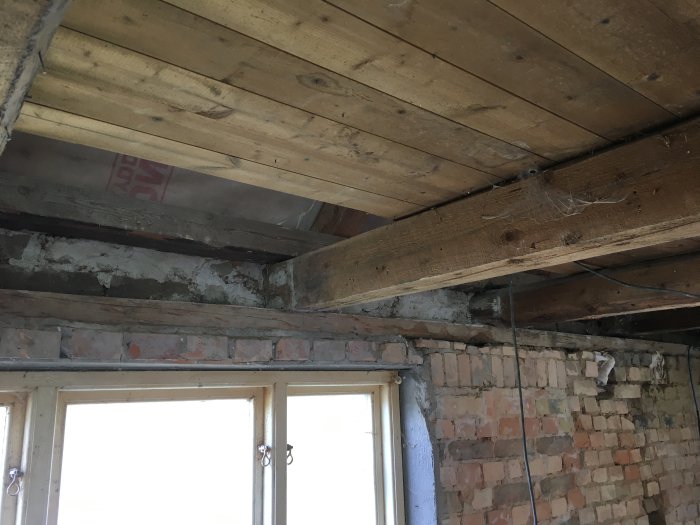 Renovering pågår, träbjälkar i tak, tegelvägg, fönster, rör, spindelnät, ofärdigt rum, konstruktionselement synliga.