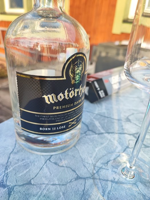 Tom whiskyflaska med märket "Motörhead" samt ett tomt glas på ett blått bord utomhus.